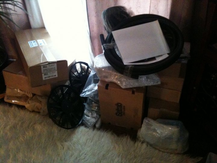 Santa Bring Pistonheads - The image captures a cluttered room corner with various items piled high. Dominating the scene is a large cardboard box on the right, possibly containing items such as a tire, a fan, and other miscellaneous objects. A computer monitor is also present, adding to the chaotic environment. In the foreground, a white furry blanket partially covers the ground, contrasting with the worn-down rug where these items rest. The space is dimly lit, with a hint of natural light coming in from the window on the left. The room corner appears to be a garage or storage area, given the disarray.