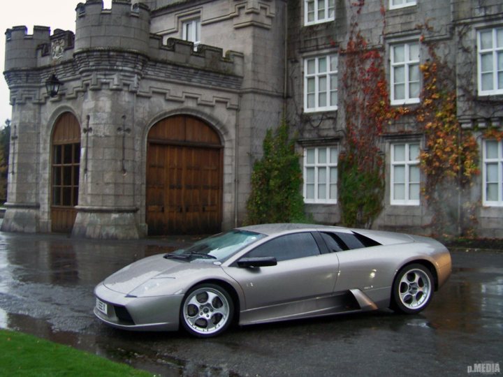 Clannonball Pistonheads - The image showcases a gray, two-door sports car parked prominently in front of a castle-like structure with a round sentry box next to a grand wooden door. The car is positioned on a rain-soaked pavement, reflecting its sleek design and silver color. Surrounding the building, ivy vines decorate the facade, adding a touch of nature to the scene. The sky overhead is overcast, creating a somewhat dramatic backdrop for the car and the castle.