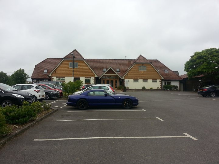 A car parked on the side of the road - Pistonheads - The image depicts an outdoor parking lot with a building in the background. In the foreground, a blue convertible sports car is parked on an asphalt surface, adjacent to a marked parking space. Several other cars, including a few with older models, are also parked nearby, indicating a public parking area. The building in the background appears to be a two-story structure with a pitched roof, possibly a lodge or a similar type of accommodation. Due to the cloudy weather, the lighting suggests an overcast day.