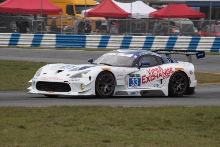 A car is parked on the side of the road - Pistonheads - This image captures an intense moment on a racetrack. A white sports car with red lettering and a large rear wing is in the foreground, driving on the racetrack with its driver focused on the road ahead. The car number is clearly visible, indicating it's a racing event. In the background, there's a fence and tents, likely set up for the event. Beyond the fence, other vehicles and people are visible, suggesting a bustling scene at the racetrack.