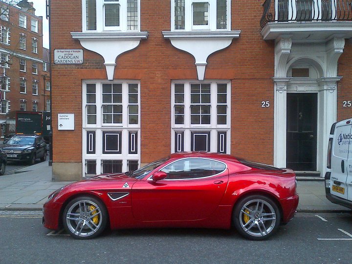 - Page 388 - Supercar General - PistonHeads - The image shows a vibrant red sports car parked on the street by the side of a brick building. The building features bay windows outlined in white, each adorned with black grids and panes. To the right of the frame, a partially obscured white van is parked. A red "25" sign is visible on the building, indicating that this might be a designated parking or zone for this address. The scene captures a quiet day on a residential street.