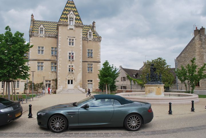 Pistonheads trip to Monaco Historique - Page 2 - Aston Martin - PistonHeads - A dark-colored luxury car is parked on the curb next to a historic, possibly European, stone building with a conical roof. The image suggests the contrast between the modern vehicle and the old architecture. People are scattered along the sidewalk in front of the building, conveying a sense of urban activity. The sky is overcast, providing a diffuse light over the scene. The bench on the left side of the frame is empty, adding to the quiet ambiance of the setting.