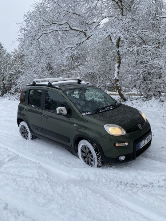 Buying Blind Pandamonium – Fiat Panda 4x4 Cross - Page 2 - Readers' Cars - PistonHeads UK - The image shows a snowy outdoor scene where a compact SUV is driving on what appears to be a partially cleared path. The vehicle has a greenish-brown body color, and there's some visible snow accumulation on the car, particularly on the roof. In the background, there are trees with bare branches indicating a winter season. The sky is overcast, further suggesting cold weather conditions.