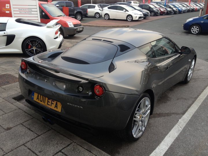 Evora 2+2 IPS deposit paid - Page 1 - Evora - PistonHeads - In the image, a sleek, black sports car is the centerpiece, parked on a street lined with various other vehicles. The sports car is ready to be driven, with its shiny exterior and silver rims reflecting the ambient light. The license plate is clearly visible, adding a personal touch to the vehicle. In the background, white cars and a large delivery truck are also parked, suggesting a busy street or a commercial area. The street itself is paved with asphalt, contributing to the urban setting. The overall scene conveys a sense of contemporary living and mobility.