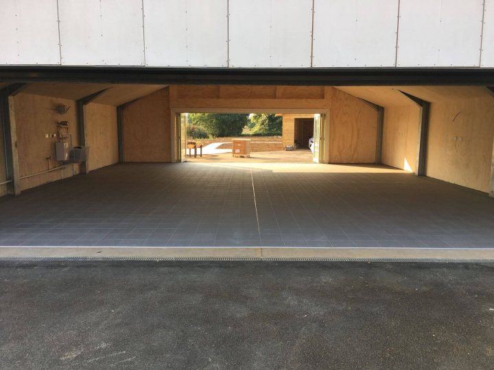 Who has the best Garage on Pistonheads???? - Page 292 - General Gassing - PistonHeads - The image depicts an empty garage or storage room. The walls and floor are constructed of concrete, and there is a garage door on the right side, which is currently closed. In the background, through a window, it appears to be daylight outside, and a clear sky is visible. There is no signage or personal items in the space, giving it an unoccupied look. The lighting inside the room is artificial, likely from overhead fixtures or wall sconces.