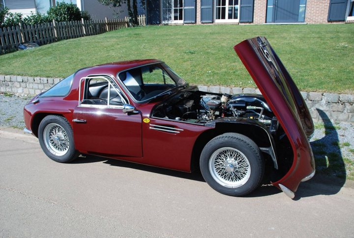 Early TVR Pictures - Page 94 - Classics - PistonHeads - The image shows an older model red convertible sports car parked on a paved area. The car's trunk and rear window are visibly open, providing a view into its interior where the engine is exposed. A brick wall is in the background, partially concealed by the grassy area adjacent to the car. The car appears to be well-maintained, but the engine work suggests it may not be road-worthy. The vehicle's design and the context it is presented in suggest a classic or vintage style.