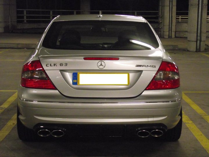 Mercedes Pistonheads - This image captures a scene in a parking lot at night, focusing on the rear of a silver Mercedes-Benz C-Class sedan. The car's license plate, featuring the alphanumeric code "CLk 63", can be read against the backdrop of the illuminated parking lot. The vehicle's left brake light is lit, indicating it is currently parked. The finishing line of the parking lot, with its distinguishable yellow and white markings, is also partially visible in the frame.