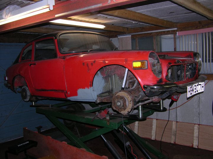 Working Moment Pistonheads - The image displays a red vintage Mercedes-Benz car being worked on in a garage setting. The car is hoisted on a lift and appears to be old and rusty, particularly around the tires and the metal framework visible beneath the hood. The interior of the garage suggests an ongoing restoration project, with the vehicle situated between wood panels and other tools required for such a task. The focus is on the car, capturing its classic design and the restoration process.