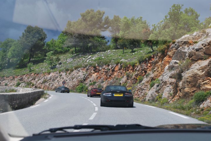 Pistonheads trip to Monaco Historique - Page 2 - Aston Martin - PistonHeads - The image shows a narrow winding road built on the side of a rocky mountain. Several cars are driving along the curve, with one prominently in the foreground while others appear further down the road. The road is lined with grassy areas and trees, and there's a large amount of stone and vegetation surrounding it. The scene suggests a scenic mountain drive, with changing landscape and varying levels of sunlight indicating that the photo was taken during the day.