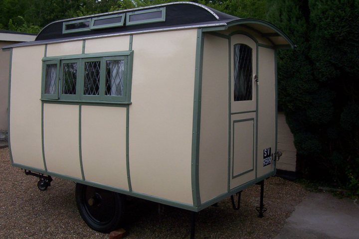 New oldies - Page 1 - Classic Cars and Yesterday's Heroes - PistonHeads - The image features a quaint, single-occupant caravan parked on a gravel driveway. The caravan has a beige exterior with a green trim around its windows and black outline framing its body. There are three small windows on the front and sidewall. The door is secured with a screen, eliminating the need for a bunch of pesky keys. The camping caravan is positioned adjacent to a building and a lush green hedge, suggesting it might be part of a backyard or a storage lot.