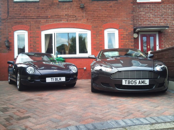 Pistonheads - The image showcases two classic cars parked side by side on a red brick driveway. Both vehicles are convertible models with a distinctive rounded shape, reminiscent of the 1960s or 1970s style. The car on the right is plain black with a plate that reads "TBO5 AMQ," while the car on the left is black with a distinctive solar panel attached to its roof and a plate that reads "TBLK." The driveway is flanked by mature trees and situated in front of a red brick house with a white-trimmed window. The overall atmosphere is peaceful and timeless.