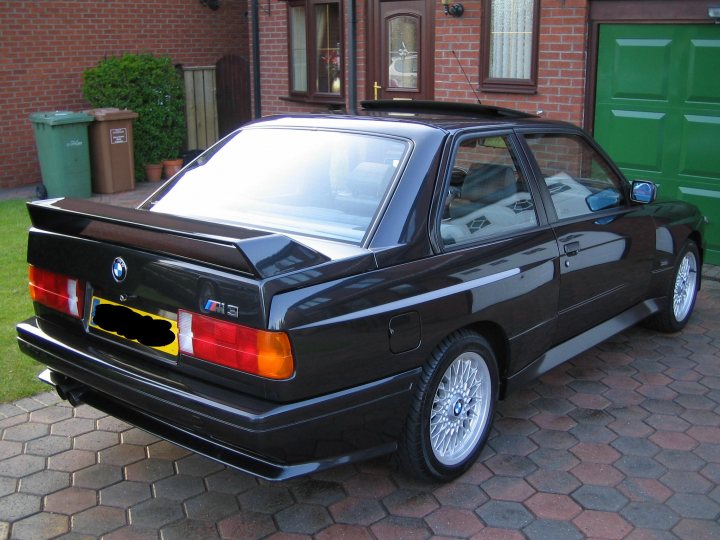 Pistonheads Contribution - The image depicts a two-door black BMW car parked in front of a green door with rectangular glass panes. The car has a distinctive sloping rear window, flared rear fenders, and a spoiler on the trunk lid. The license plate is visible, bearing the BMW logo, but the registration number should not be read here. The car's wheels are also visible, featuring a multi-spoke design. There is a small hedge to the left of the car, and a trash bin further to the left in the background. The brick driveway where the vehicle is parked sinks slightly in front of its front tire, suggesting age or dampness.