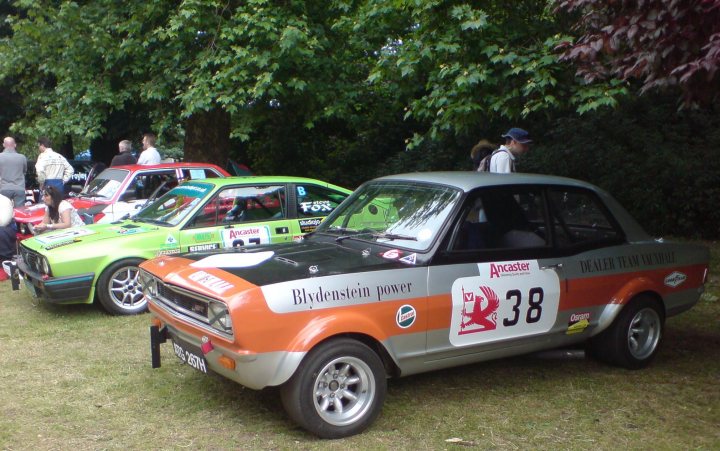 Pistonheads - The image showcases a vibrant gathering in a park, where an orange, black, and gray retro-styled car and a lime green and black classic car are parked on a grassy area with a prominent tree in the background. Both cars are exhibiting a sense of history and charm, attracting the attention of several people who are walking around, admiring the vehicles. The scene is lively and informal, suggesting a community event or a meetup of classic car enthusiasts. The cars are placed next to each other, emphasizing the vintage aesthetic and fostering a sense of camaraderie among the attendees.