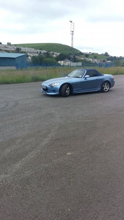 my honda s - Page 1 - Readers' Cars - PistonHeads - The image shows an open road with a blue sports car driving on the surface. The car is captured from a rear three-quarter angle, emphasizing its sleek design and two-door coupe configuration. The sky above is overcast, and there are rolling hills in the distance, suggesting a rural or semi-rural setting. There is no text visible in the image, and no people or significant objects in the immediate vicinity of the vehicle, highlighting the vehicle itself as the main subject of the photograph.