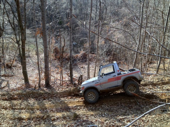 Bit of off-roading, and a bit of camping. - Page 1 - Off Road - PistonHeads - The image captures a scene in a wooded area. Dominating the frame is a four-wheeled vehicle, possibly an ATV, parked in the middle of the scene. The vehicle appears to be a darker color and is firmly planted on a trail that cuts through the woods. The surrounding landscape is a tapestry of trees and foliage, with their earthy hues suggesting a natural environment. A dirt path can be seen running parallel to the vehicle's path, inviting curiosity about the destination it leads to. From the perspective of the image, it seems to be taken from a slightly elevated viewpoint, giving a sense of the scene and offering a glimpse of the vehicle's placement within the woodland.