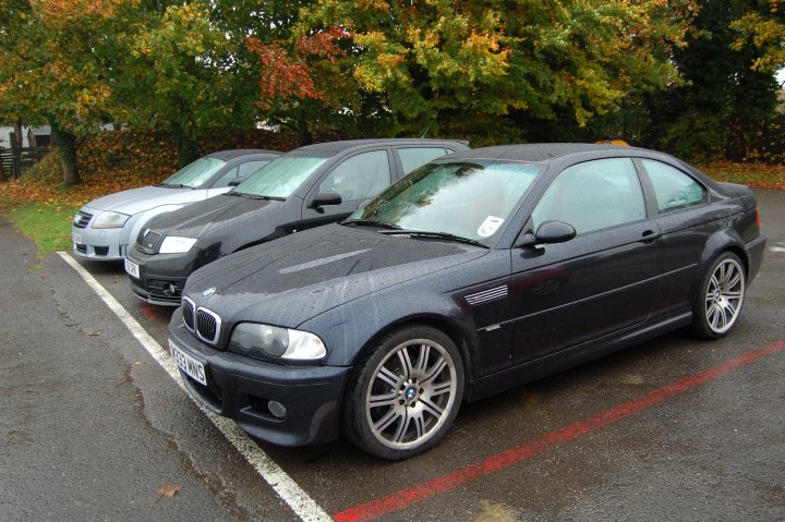 Pistonheads - This image depicts three cars parked in a lot with a red painted line marking the edges of the parking spaces. The car in the foreground is a dark blue BMW with white logos, polished nickel grille, and distinctive multi-spoke wheels. The other two cars, positioned behind it, are much broader and appear to be compact sedans with black paint and silver accents, suggesting they might be from a different brand. The ground is wet, which could indicate recent rainfall, and there are trees in the background, indicating that the lot is fairly large or set within a park-like environment.