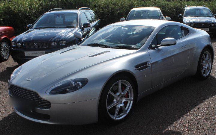 Foray Foreign Pistonheads Belated - The image shows a setting with a group of luxury cars. A striking feature is a silver Aston Martin with its hood up, which is prominently positioned in the foreground. The Aston Martin is parked on a gravel drive with other cars around it. The texture of the gravel and the cars' sleek designs are notable details in this scene.