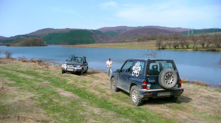 background car pics :) - Page 15 - Readers' Cars - PistonHeads - The image captures a peaceful countryside scene. There are two cars parked along the edge of a body of water, possibly a lake. A person is standing between the two cars, overlooking the water. The cars are adorned with stickers and appear to be old models, possibly with off-road capabilities given their rugged look. The water is calm, reflecting the serenity of the scene.