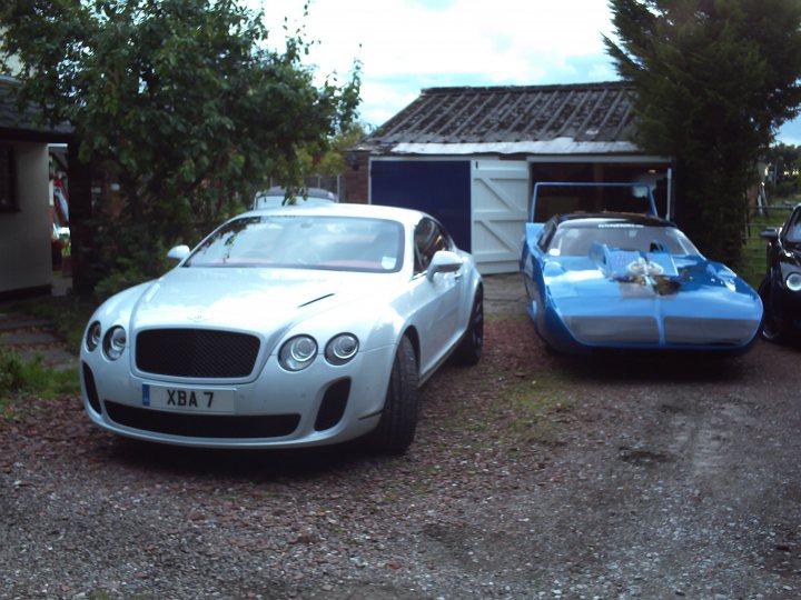 Supersports - how long before depreciation levels out - Page 1 - Rolls Royce & Bentley - PistonHeads - This image depicts a gravel driveway where two vintage-style cars are parked. On the left side, there is a white Bentley XBA with a distinctive grille and LED headlights. On the right side, a blue convertible is parked under the shade of a tree. Both cars appear to be luxury models, with the Bentley being particularly distinctive due to its iconic design. The setting includes a garage and a tree with lush green leaves, indicating a well-maintained residential area. The overall tone of the image is one of luxury and classic car design.