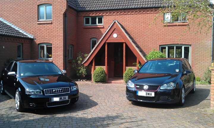 Who drives what? - Page 1 - East Anglia - PistonHeads - The image shows an outdoor scene with two black cars parked on the brick driveway of a two-story brick house. The house features a peaked roof and prominent front door, and there is a garage with an open door visible in the background. The cars appear to be modern, and one of them displays a European license plate. The setting suggests a residential area, and the presence of vehicles implies that the image was captured during the day. Overall, the image captures a quiet, pleasant day in a suburban neighborhood.