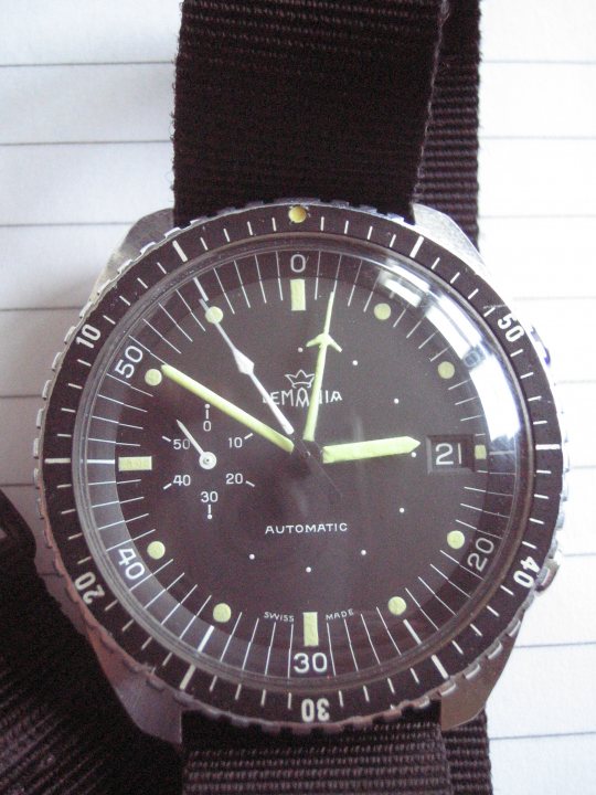 Air Issue Pistonheads South Lemania Force African - The image displays a wristwatch with a black face and golden numbers and markings. It appears to be a diver's watch with a timer layout and luminescent hands and hour markers. The watch is secured with a black strap and a piece of paper or fabric is partially visible in the background. The style of the watch suggests it might be vintage or a reproduction of an analog timepiece, common among divers or casual wear styles. The image is a close-up with the watch focused in the foreground, providing a detailed view of its features.