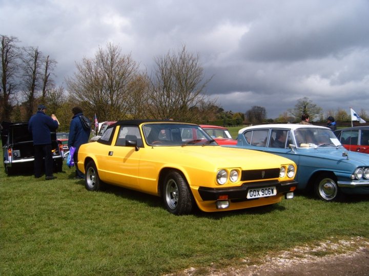 Classic Pistonheads Tatton Park - The image presents a gathering of vintage cars in a grassy field, under a gloomy sky. There's a bright yellow Triumph GT8 parked prominently in front of two other cars, the majority of which are blue. Several people are scattered around, some closer to the cars than others, suggesting a casual, open-air environment. The overall atmosphere is overcast, hearkening back to an era when such cars would have been actively used.