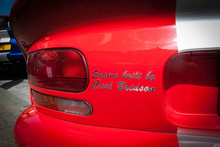 A red truck is parked in a parking lot - Pistonheads - The image features the rear section of a vibrant red car with a glossy finish. The tail light, which is the central focus, displays a message "Engine built by Carl Bridson". This indicates a personalization, accentuating the car's character. The license plate is not visible in the image. The background, though blurred, suggests the setting to be an outdoor parking area. The overall scene conveys a sense of individuality and pride in the car's craftsmanship.