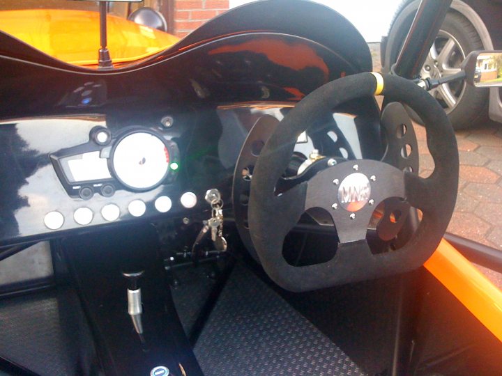 Pistonheads Contribution - The image presents a close-up view of a car steering wheel. The wheel is well-maintained, featuring a black leather upholstery with the infinity logo prominently displayed in the center. It's attached to a black console that houses numerous controls and buttons. The background subtly shows a glimpse of a street scene. The style of the image is a realistic photograph, focusing on the car's interior.