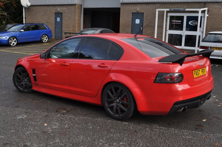 Pistonheads - The image depicts a bright red, two-door coupe parked in a parking lot. The car is adorned with a black racing spoiler on the rear, which adds a sporty touch to its appearance. It's a neatly maintained vehicle, featuring five-spoke black rims that complement its vibrant exterior. The car is set against a backdrop of a brick building with a white frame door and several cars parked, suggesting a common parking area, possibly belonging to the workplace or a public parking zone.