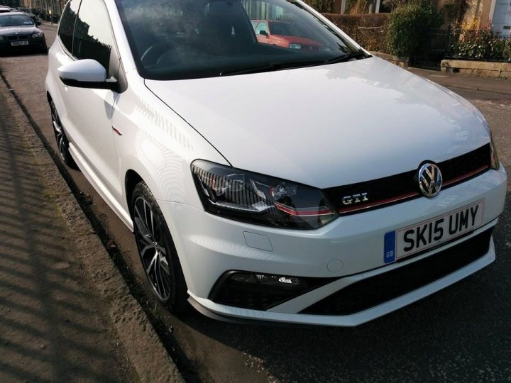 A white car parked next to a parking meter - Pistonheads - The image shows a white Volkswagen GTI parked along the side of a road. The car features black rims and a red accent line, with a "GTI" badge prominently displayed on the side. The license plate reads "SK15 UMY." The vehicle is shiny and well-maintained, reflecting its status as a premium model. In the background, there is another car, partially visible, and a fence, suggesting a residential or commercial area. The image appears to be taken during daylight, with natural light illuminating the scene.