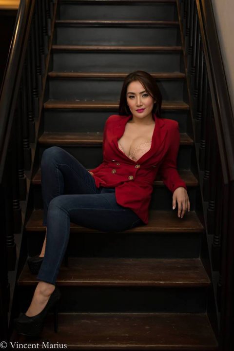 A woman sitting on a bench with her legs crossed - In the image, a woman is situated on a set of indoor stairs. She is stylishly dressed in red clothing, including a red coat. One foot is playfully placed on the stair, adding a unique pose to the scene. The woman is looking upwards with a smile, giving the image a sense of warmth and approachability.