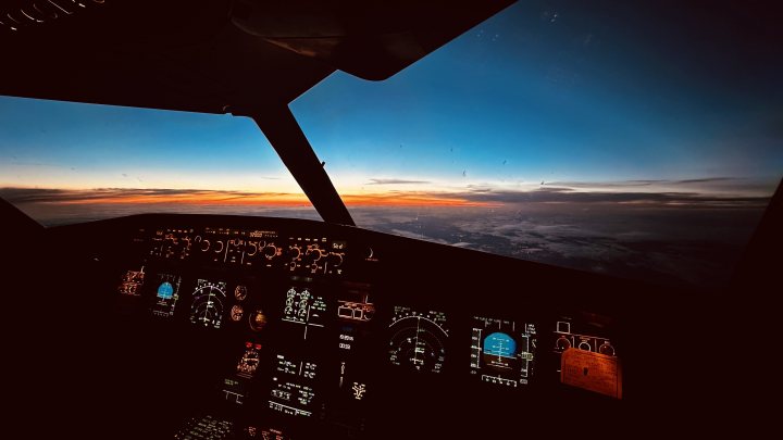 The image shows the interior of a small airplane cockpit at what appears to be sunrise or sunset, given the warm hues in the sky. The view is from the back seat towards the front, providing a clear view of the dashboard and instruments. The cockpit is well-lit, indicating that the photo was taken during daylight hours with natural light entering through the windows. The overall scene suggests a calm and routine moment before or after a flight.