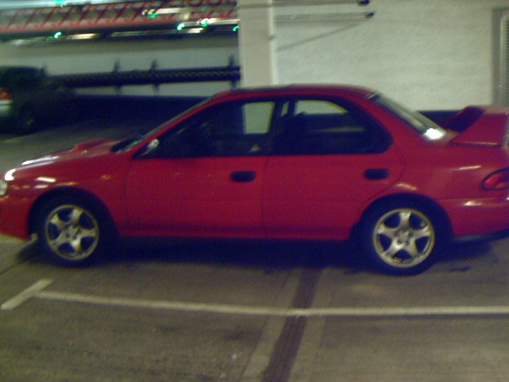 Impreza Pistonheads Turbo - The image depicts a red coupe parked in a public parking lot during night or early evening. The car has a noticeable body kit and has a spoiler mounted at the rear. Its color matches the parking lot lines, enhancing the visibility of the lines. The surrounding environment is relatively dark, emphasizing the red car and the white lines.