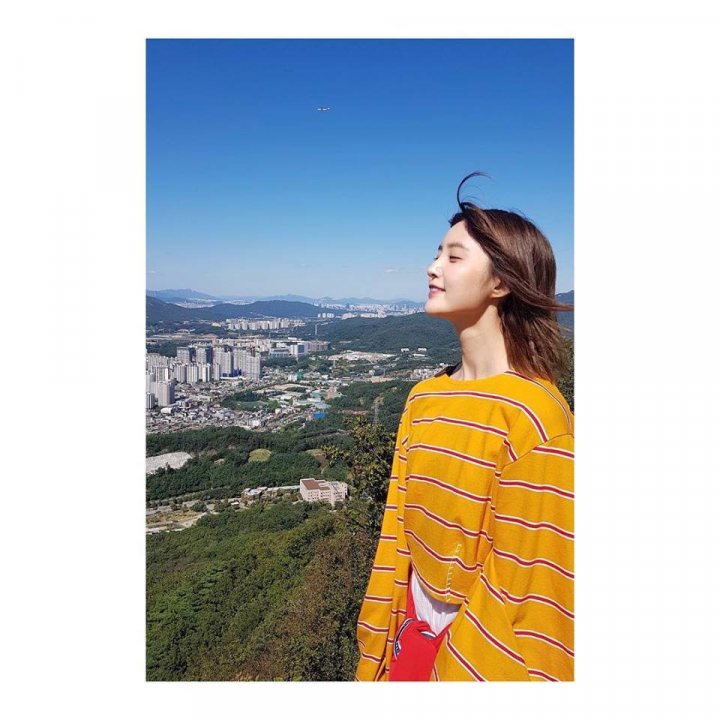 A woman standing in a field holding a kite - The image shows a young woman standing outdoors, gazing upwards into a clear blue sky. She's wearing a vibrant yellow striped shirt and has her hair pulled back. Below her, there's a sprawling urban landscape with concrete structures and a sea of trees, clustering towards the center. Above, an airplane is visible, adding a sense of scale and depth to the scene. The woman's face is lit up with a relaxed or content expression, suggesting she's enjoying the view.