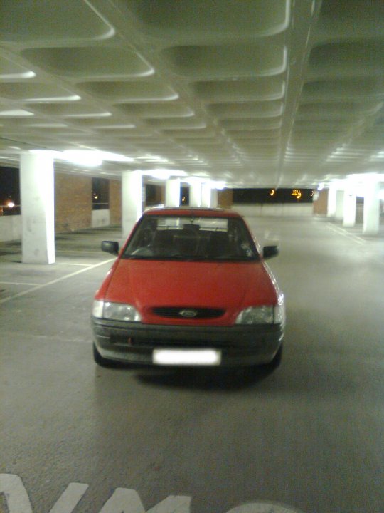 Escort Pistonheads - The image features an abandoned parking garage. In the center, there's a small red car, parked and facing the camera. The car has a distinctive square theme that continues with the white ceiling of the garage. There are no other vehicles or people visible in the scene, adding to the sense of abandonment. The garage's structure is quite simple, with white concrete pillars and a black vent at the back. The overall atmosphere suggests the photo was taken during dawn or dusk, as the lighting is soft and muted.