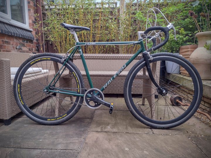 The "Show off your bike" thread! (Vol 2) - Page 88 - Pedal Powered - PistonHeads UK - The image shows a vintage bicycle leaning against a patio table. It features a distinctive color scheme with red, white, and green detailing. There are also black and yellow wheel decals visible on the wheels. A wooden fence is in the background, suggesting an outdoor setting, possibly a garden or backyard. The style of the bicycle, with its retro design, suggests it may be a collector's item or used for special occasions like vintage cycling events.