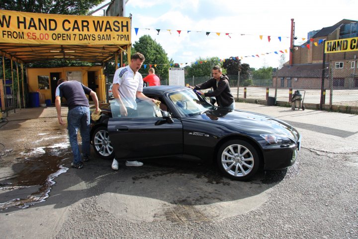 My S2000 review - Page 5 - Honda - PistonHeads - The image depicts an outdoor scene involving a black car being washed. Two individuals are actively engaged in the wash process; one person is rinsing, while the other is lathering a cloth to apply soap on the car's surface. The car is parked on a paved surface, possibly a parking lot. A banner hanging above the workers advertises a "HAND CAR WASH." Additional flag bunting decorates the area, indicating a festive or promotional event. The background includes a building with a parked bicycle in front of it, suggesting this car wash could be a part of a larger facility or complex.