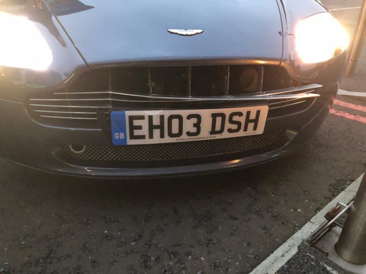 Vantage Grille - Page 2 - Aston Martin - PistonHeads UK - The image shows a scene on a road with a dark-colored car in the foreground. The car has a broken front bumper, which is hanging off the vehicle. A white license plate that reads "E00 3SH" is visible on the back of the car. The car's design features distinctive grille patterns and what appears to be an emblem that could indicate the make or brand. The surroundings suggest it might be a street, and there are no people or other vehicles immediately around the car in the image. The focus is on the car itself, which is centered and occupies most of the frame.