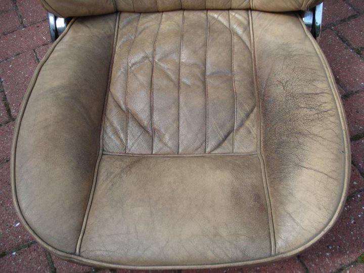 DIY Leather restoration - My weekend experience (Pictures) - Page 1 - Classic Cars and Yesterday's Heroes - PistonHeads - The image features an outdoor reclining chair that appears to be made of leather. The chair is situated on a plain, light-colored ground surface that seems to be a paving area or similar outdoor setting. The leather material on the chair shows signs of wear, with noticeable creases and stains, particularly on the backrest and seat cushion. The chair is empty and there is no distinguishable text or markings on it that could provide additional context or information.