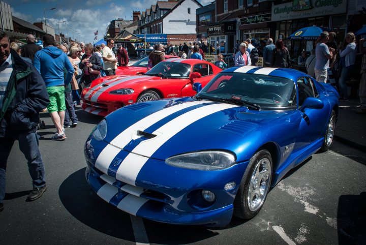 A car is parked on the side of the road - Pistonheads - This image captures a vibrant street scene, featuring a pair of striking sports cars parked on the side of the road. The cars are bright and polished, with a design that includes a white stripe contrasting against the blue body. People are milling around the scene, adding a lively atmosphere to the image. Buildings with a mix of architectural styles create a diverse urban backdrop. The overall image exudes a sense of excitement and anticipation, perhaps indicating the beginning of a racing event.