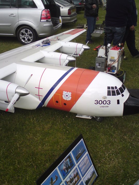 Bomber Pistonheads Kemble Vulcan - This image captures a lively scene on a grassy field under a clear sky. The main focus is a large white model airplane with orange and blue stripes that extends across most of the image. Scattered around the airplane are two stationary cars, one of which has its trunk open. There are also several people in the background, and two cars are visible further back in the field. The overall atmosphere suggests an outdoor event or gathering, possibly related to model airplanes or an aviation-related occasion.