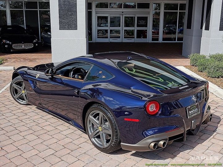 A blue car parked next to a red fire hydrant - Pistonheads - The image shows a bright blue sports car parked at the side of a building. The car is a 2019 Ferrari GTB, identifiable by its distinctive aerodynamics and the prancing horse logo on the front. It features the classic Ferrari design with a long hood and a short rear wing. The vehicle's bodywork is sleek, with accents and air vents designed to help manage airflow for optimal performance. The car is parked on a brick surface under the shade of a tree, with the backdrop of the building that appears to be a commercial or store building.