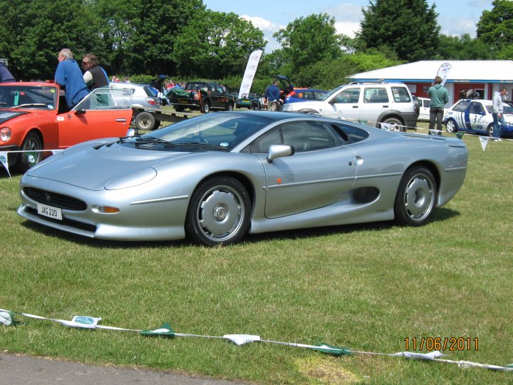 Classic & Sports Car Action Day C/Coombe.11th June - Page 1 - South West - PistonHeads - The image captures an outdoor gathering with several cars parked on a grassy area. At the center of the composition is a striking, metallic blue-gray convertible sports car. Accompanying the sports car in the scene are a variety of other cars, suggesting a car show or similar event. People can be seen milling about the area, likely admiring the vehicles or socializing. In the background, a line of orange cones marks off an area, perhaps indicating a boundary or guiding the flow of the event.