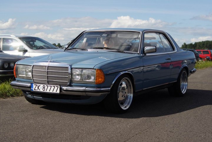 A car parked in a parking lot next to a car - Pistonheads - The image shows a classic car parked on a gravel road, with a focus on its front profile. The vehicle is a Mercedes-Benz, identifiable by its emblem and distinctive grill. It has a blue body, a color that complements the chrome grill and wire wheels, adding to its vintage charm. The car is positioned centrally in the frame, with a second car visible in the background, though it's out of focus. The sky above is clear, suggesting a bright day. No visible text is discernable in the image.