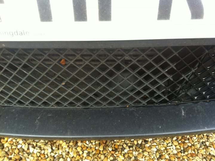 Unidentified Speed camera detector fitted in front grill - Page 1 - General Gassing - PistonHeads - The image shows a close-up view of a license plate protector, featuring a black pattern and metal grid design. Only a small portion of the license plate is partially visible through the slats in the grill. Below the protector, there is a gravel ground which extends across the frame of the photo. The focus of the image is on the license plate area, with the background being out of focus. No text or numbers are discernible in the image.