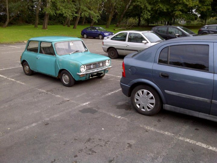 Newly Acquired Early Clubman - Page 1 - Classic Minis - PistonHeads - This image presents an outdoor parking area with a variety of cars, among which a teal-colored vintage car stands out due to its bright color and distinct style compared to modern vehicles. The vintage car is parked on the left, between two more contemporary cars, showing a contrast in design and era. The parking lot itself is large and paved, with a few other vehicles parked throughout the area under various degrees of shade. The background suggests the presence of trees and grass, indicating a relatively park-like environment or a green zone. The weather appears to be sunny, possibly during the day.