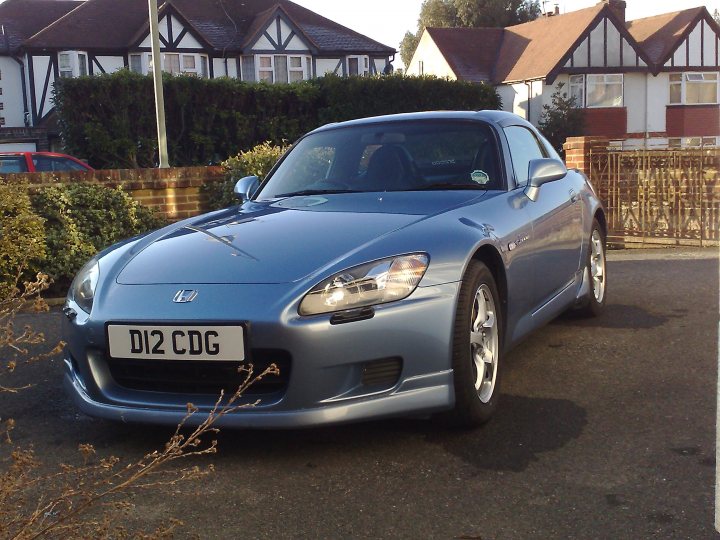 Stand Blue Light Pistonheads Touchpaper Nurburgring - The image shows a blue Honda sports car parked on a paved road. The vehicle is positioned at an angle to the camera, highlighting its front end and profile. The surroundings include a house with a white facade and black roof, a brick wall, and some greenery. The Honda emblem is prominently displayed on the front of the car.