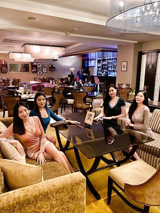 A group of people sitting around a table - In the image, there is a spacious and modern restaurant interior with high ceilings. The floor is carpeted in beige, and there's a large chandelier hanging from the ceiling. Four individuals are seated around a wooden dining table, which is set for three people. They are engaged in conversation and enjoying their time together. The walls of the restaurant are adorned with framed pictures that add a personal touch to the space. There's also a bar counter visible in the background. Overall, it appears to be an inviting atmosphere suitable for socializing and dining.