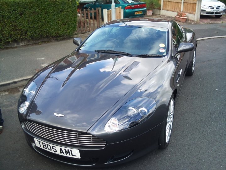 Have to tell someone - Page 1 - Aston Martin - PistonHeads - The image features a black sports car with a shiny metal finish parked on the street. It has a sleek, modern design with alloy wheels and a prominent grille with a car logo. The sky reflects in the car's polished surface, suggesting that it has been rained on recently. In the background, there are other vehicles parked along the side of the road, and a wooden fence can be seen. The car takes up a significant portion of the image, indicating its prominence in the scene.