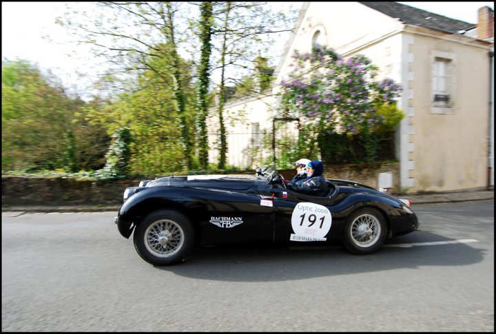 The Tour Auto came to town - Page 1 - Classic Cars and Yesterday's Heroes - PistonHeads - The image depicts a vintage black car in motion on a road. The car is driving towards the camera and bears a number 191, which could suggest it's participating in a classic car race or event. Surrounding the car are lush green trees, hinting at a rural or semi-rural setting. There's an indication of a highway or main road, as there's a discussion of whether this is the M 22 in Italy. The car and its two occupants are the primary subjects, with the driver and passenger clearly visible inside.